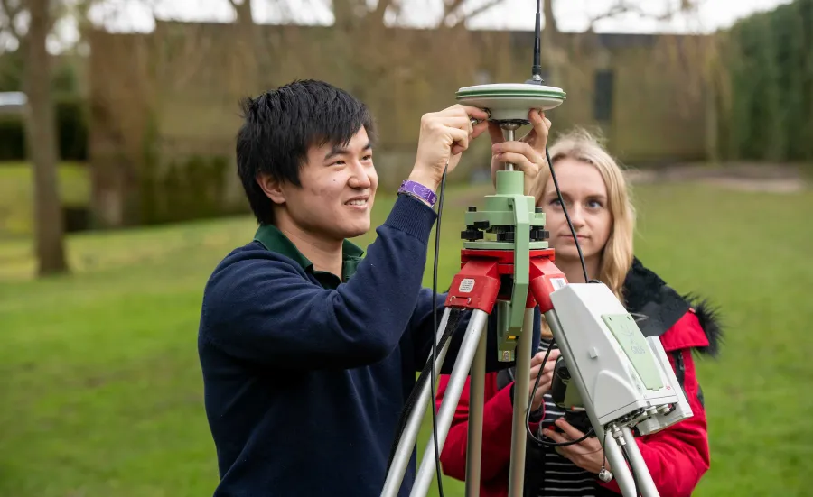 Two students conduct an experiment outdoors using a large piece of survey equipment on a tripod.