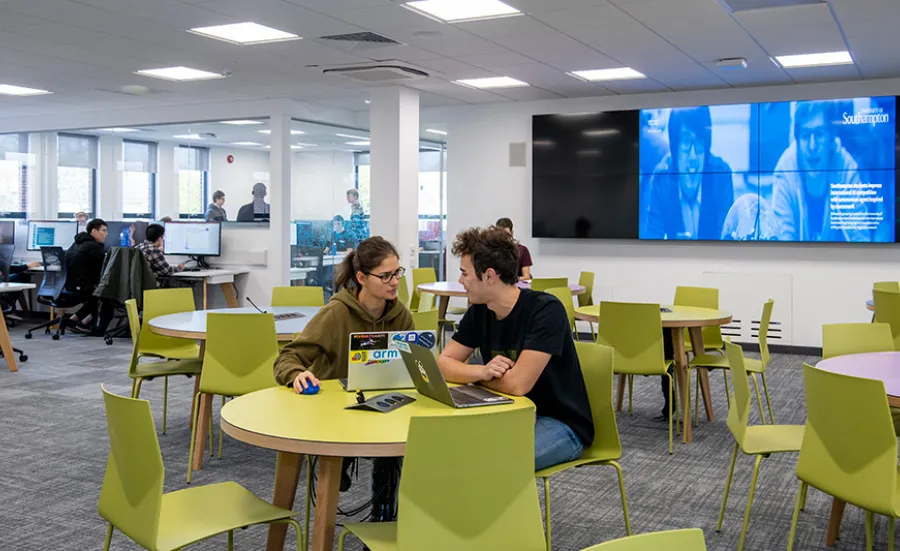 A wide shot of the software projects laboratory, with two students discussing a project at a table in the foreground, and more students working at computers and in a conference room further behind.