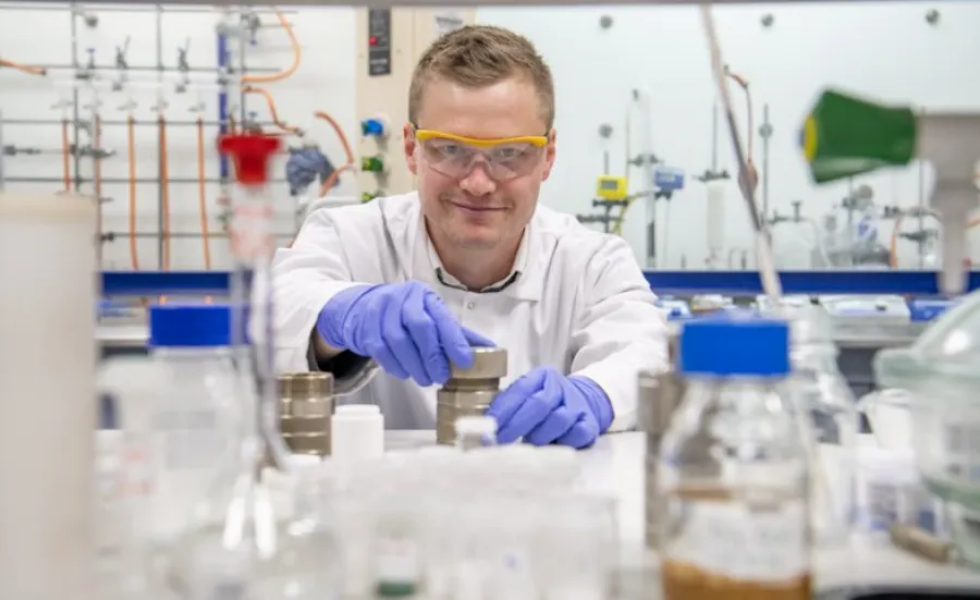 A chemistry researcher in goggles, gloves and white coat works with equipment in the lab