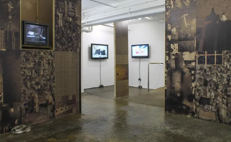 The Taplow House art installation in ASC Gallery. The installation walls in the foreground are covered in dark, industrial images. In the background, they are white. Several televisions hang on the walls.