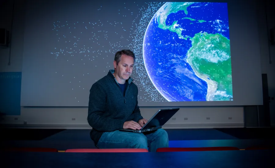 Professor Hugh Lewis sitting on the front bench of a lecture theatre as he consults his laptop. A graphic of satellites orbiting Earth is projected on the screen behind him.