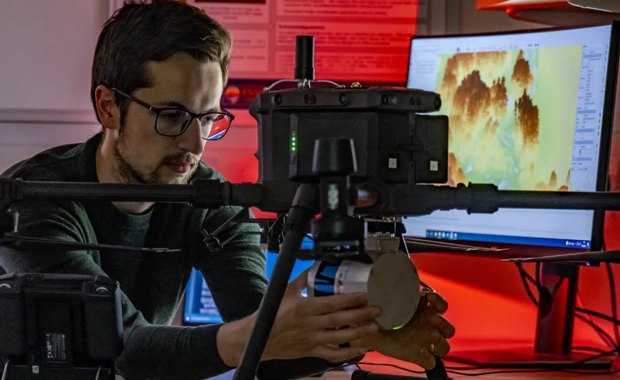 A researcher checks a remote sensing drone. To their right is a computer screen with a topographical visual