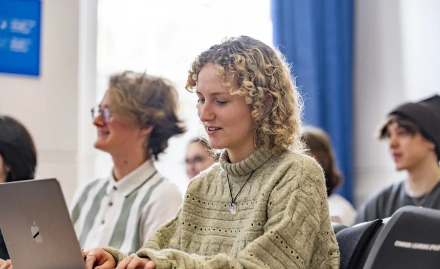 A student is sitting at a desk, typing on their laptop. They have blonde curly hair and a beige knitted jumper. Other students are sitting in the same room but are out of focus. They all face the same direction and are smiling.