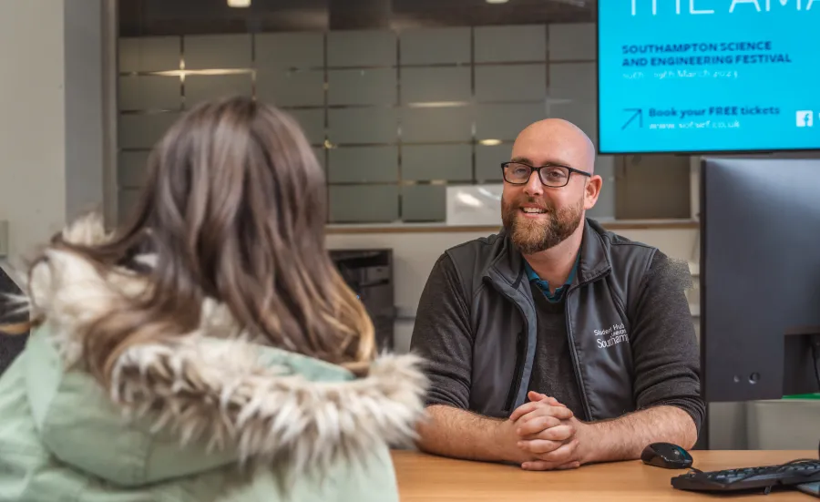 A University of ƱAPP_ĿͲƱ-ٷϷ, staff member sits behind a desk while speaking with a student. The staff member is smiling and wearing University of ƱAPP_ĿͲƱ-ٷϷ,-branded clothing.