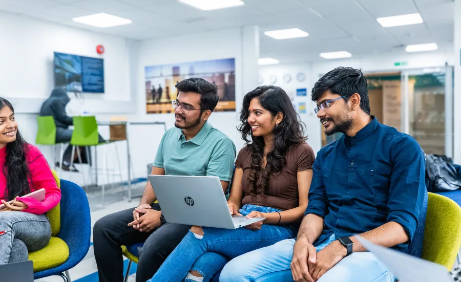 A group of students seated together in a modern study environment, discussing with laptops and notebooks, smiling and engaging in conversation