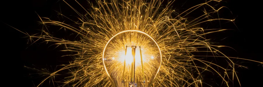 An incandescent light bulb on a reflective surface with a glowing filament surrounded by a halo of golden sparks against a dark background.