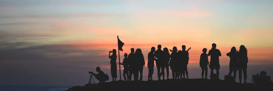 A group of people as silhouettes against a colorful sunset sky, some taking photos and holding a flag, on a rocky lookout.