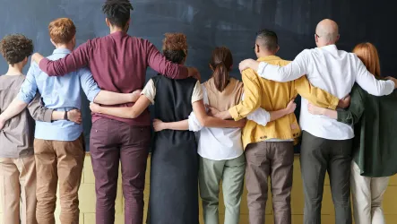 A diverse group of nine people stand in a row with their arms around each other's shoulders, facing a blackboard in a classroom, dressed in an array of casual to smart-casual attire.