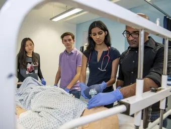 A group of students watch as one of them works on a dummy patient in a hospital bed