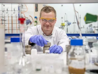 A chemistry researcher in goggles, gloves and white coat works with equipment in the lab