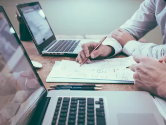 Close up view of two people working at a desk with laptops, paper and pens