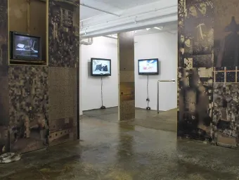 The Taplow House art installation in ASC Gallery. The installation walls in the foreground are covered in dark, industrial images. In the background, they are white. Several televisions hang on the walls.