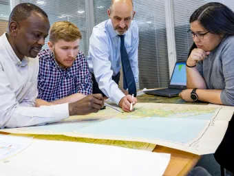 A ƱAPP_ĿͲƱ-ٷϷ, Geospatial project team meets, with one researcher standing up, pointing to a map that his colleagues are gathered around