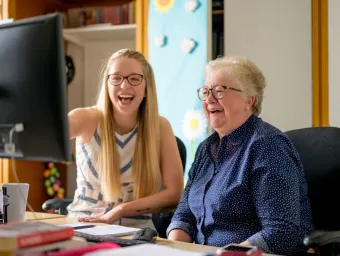 Two laughing women, one young, one older, sit in front of a computer screen. The young woman points at the screen.