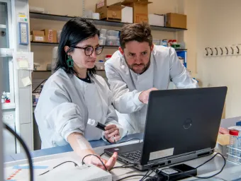 Two people in a laboratory. One is having measurements taken from their arm, the other directing their attention on a computer screen.