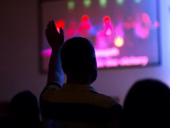 People in a room silhouetted against a screen. The image on the screen is unclear. One of the people has their arm raised to ask a question.