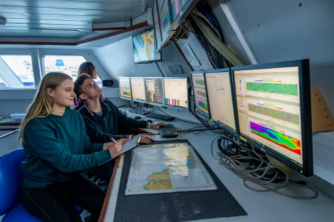 Two students sit at a desk inside a room on a boat. They observe a bank of computer screens displaying various data and information.