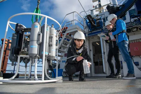 Three students outside on the deck of a boat, on a sunny day. One crouches down to collect a water sample from a large piece of technical equipment.