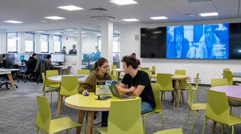 A wide shot of the software projects laboratory, with two students discussing a project at a table in the foreground, and more students working at computers and in a conference room further behind.