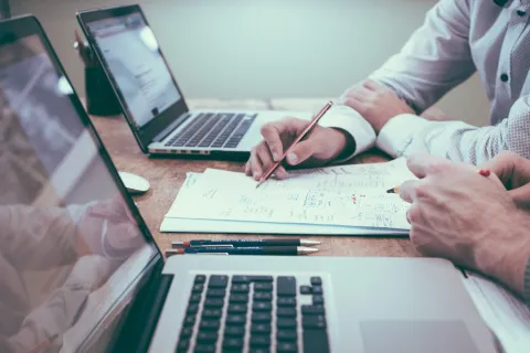 Close up view of two people working at a desk with laptops, paper and pens