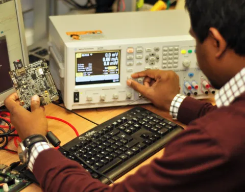 A man in a brown jumper examines a circuit board at a desk