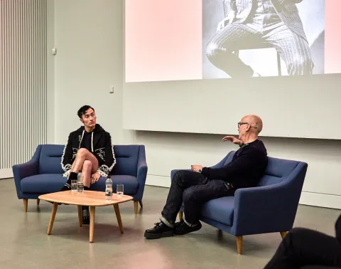 Charles Jeffrey and Professor Jonathan Faiers sit on blue sofas with a small table between them. Jonathan is facing away from the camera and has an arm raised as he speaks. Charles is facing Jonathan and appears to be concentrating on what Jonathan is saying. There is a projector behind them showing a person wearing a pinstripe suit.