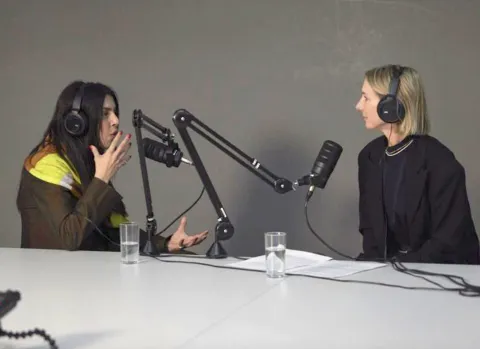 Two people sit behind a desk. Both are wearing large headphones and have large professional microphones in front of them.