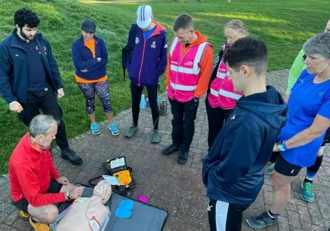 A group of people is observing a CPR training demonstration outdoors, with one person performing the demonstration on a training mannequin next to an AED device, in a sunny, grassy area.