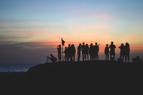 A group of people as silhouettes against a colorful sunset sky, some taking photos and holding a flag, on a rocky lookout.