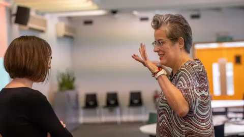 Two people are having an animated discussion. The one on the right is holding both hands up and is smiling. The one on the left has her arms folded and seems to be listening. 