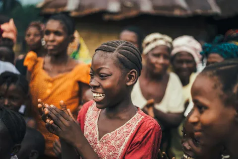 A young girl with braided hair claps and smiles amidst a group of villagers outdoors, capturing the vibrant spirit of Sierra Leone.