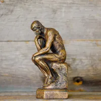 A small replica of 'The Thinker' - a bronze sculpture by Auguste Rodin depicting a man hunched over on a rock in contemplation - set against a backdrop of wood panels.