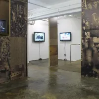 The Taplow House art installation in ASC Gallery. The installation walls in the foreground are covered in dark, industrial images. In the background, they are white. Several televisions hang on the walls.