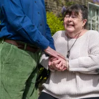 A woman in late middle age sits and holds hands of a man standing next to her