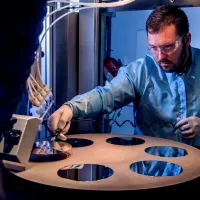 A bearded young scientist in protective overalls and glasses works with a flame hydrolysis deposition (FHD) system